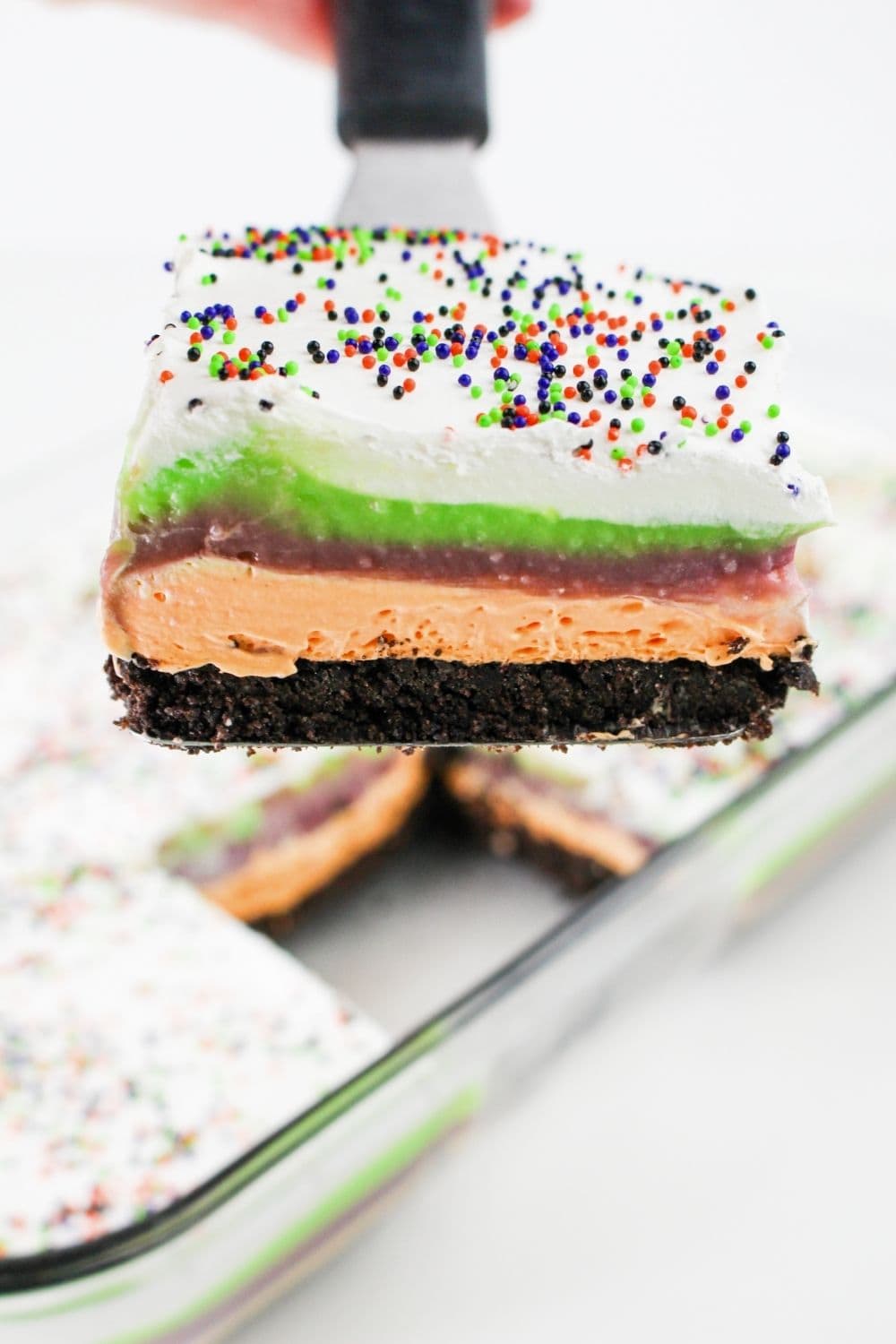 a spatula lifts out a serving of layered Halloween lush dessert, a no-bake treat made with Oreo crust, cream cheese layer, and pudding layers.