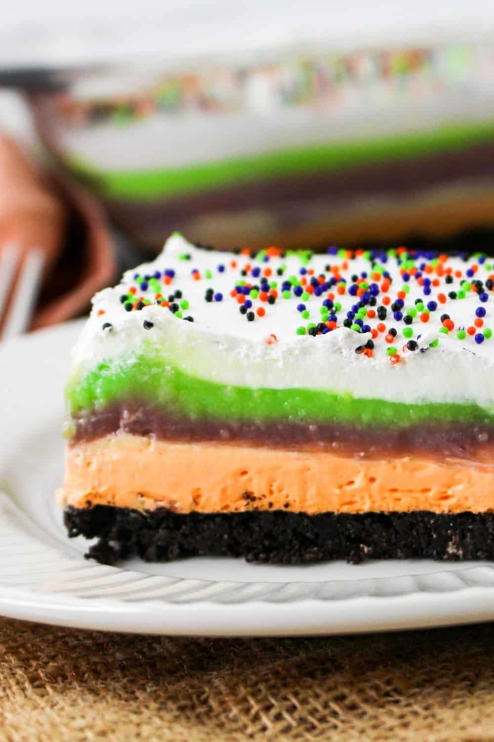 side view of a slice of Halloween layered lush dessert, showcasing the colorful layers of the Oreo crust, orange cream cheese layer, purple pudding layer, green pudding layer, and whipped cream.