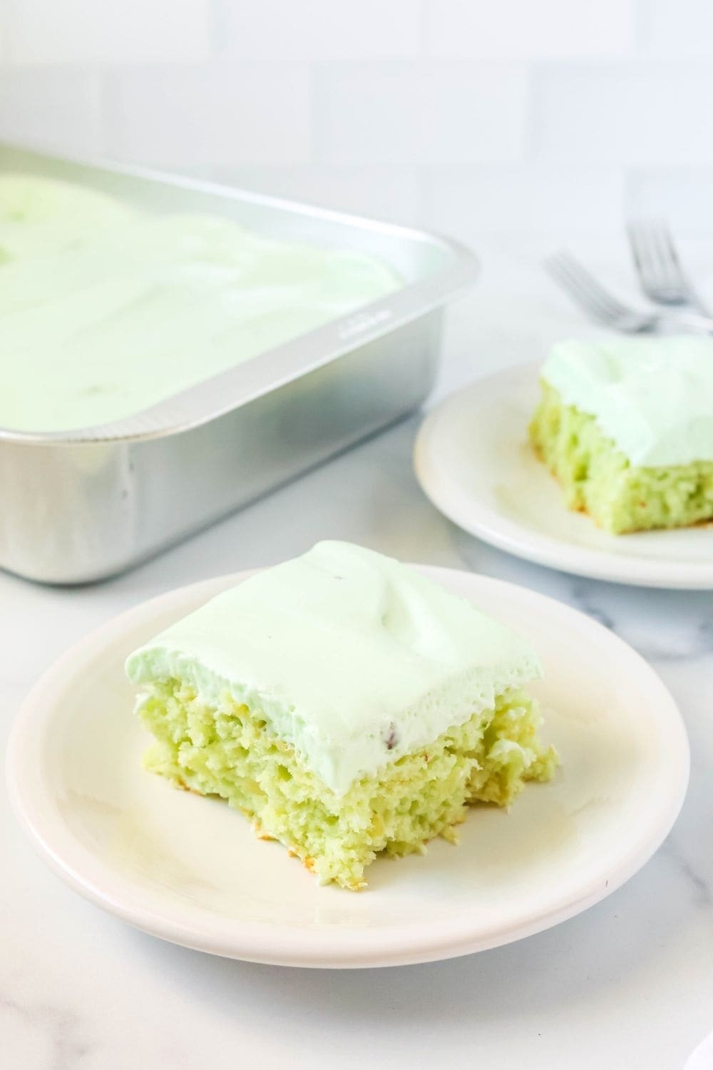 a slice of pistachio pudding and pineapple cake on a white plate, with another slice and the remaining pan of cake in the background.