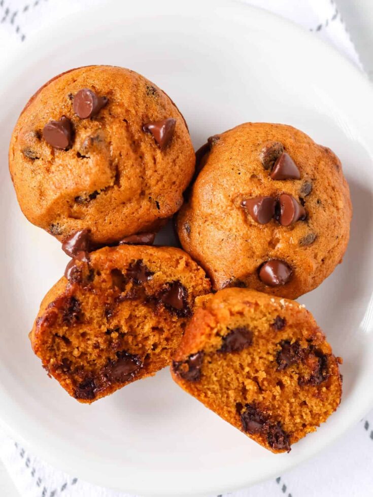 three homemade pumpkin chocolate chip muffins on a white plate. One muffin has been cut in half, showing the moist interior dotted with melty chocolate chips.