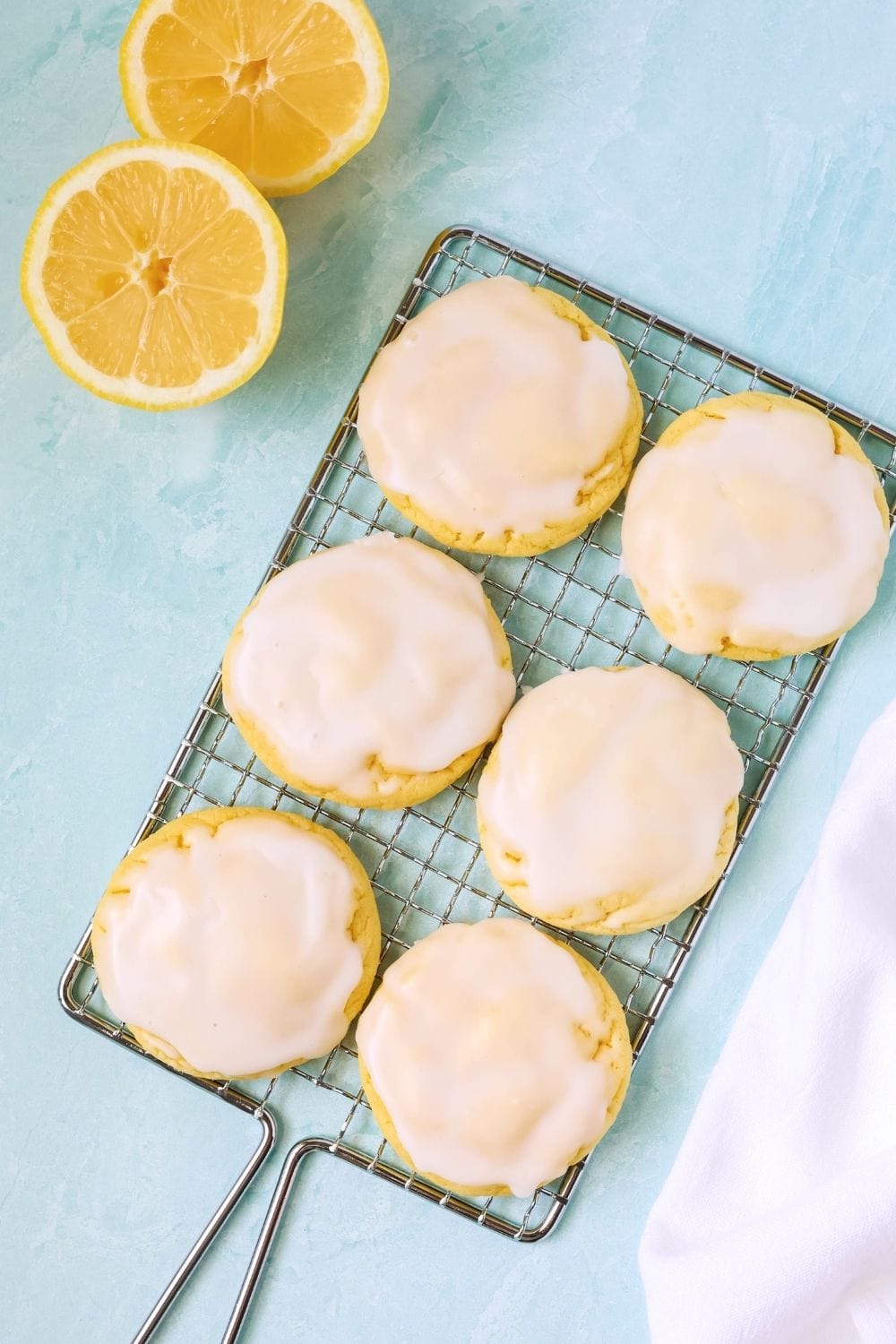 overhead view of six iced lemon cookies made with cake mix on a wire rack. Two lemon halves are next to the rack.
