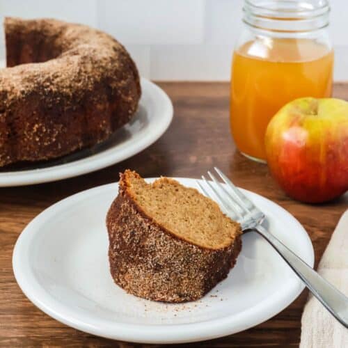 a slice of apple cider donut bundt cake from a mix served on a white plate. A glass of apple cider and an apple are on one side of the plate, and the remaining bundt cake is on the other side.