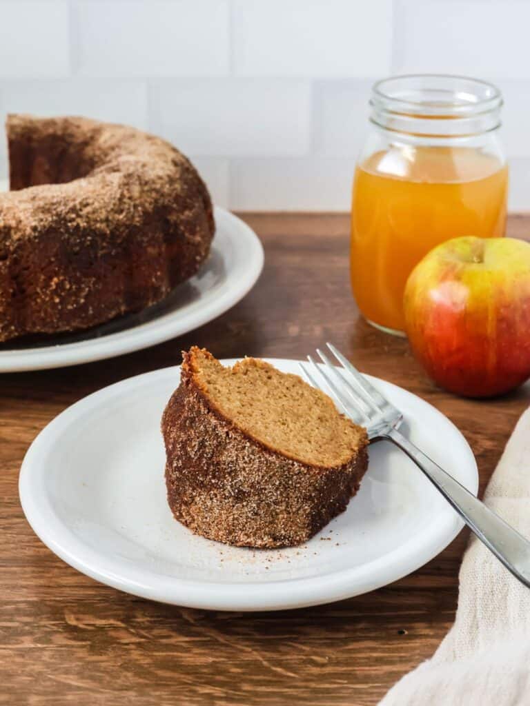 a slice of apple cider donut bundt cake from a mix served on a white plate. A glass of apple cider and an apple are on one side of the plate, and the remaining bundt cake is on the other side.