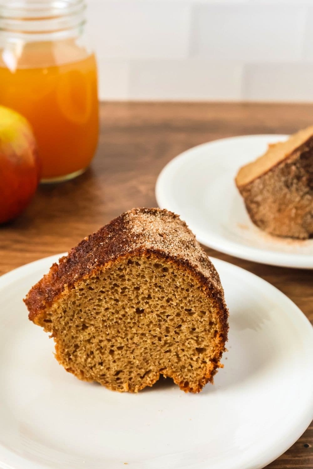 a slice of cake mix apple cider donut cake served upright on a white plate. Another slice is in the background, along with a jar of apple cider.