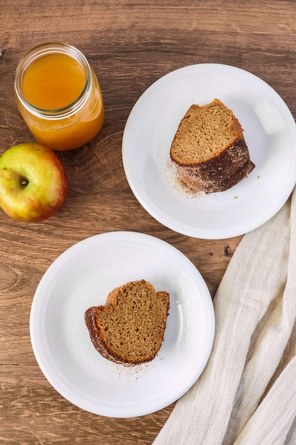 overhead view of two slices of apple cider donut bundt cake served on white plates, alongside a glass of cider and a fresh apple.