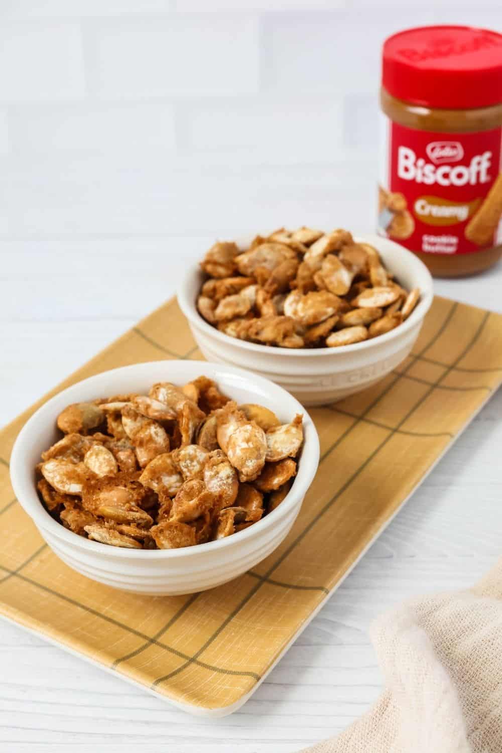 two white bowls of Biscoff-glazed pumpkin seeds, with a jar of cookie butter in the background.