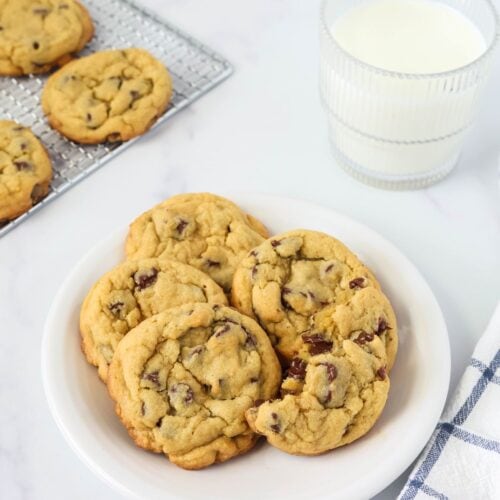 chocolate chip pudding cookies are served on a white plate, alongside a glass of milk and a wire rack of more cookies.