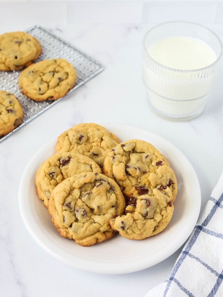 chocolate chip pudding cookies are served on a white plate, alongside a glass of milk and a wire rack of more cookies.