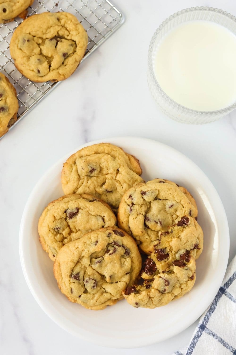 overhead view of a white plate with five chocolate chip vanilla pudding cookies on it, one of which has been torn in half. A glass of milk and cooling rack are in the background.
