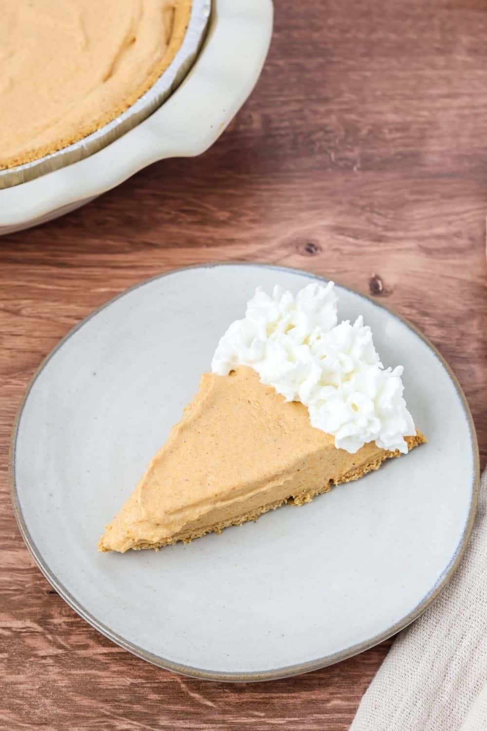 a slice of creamy pumpkin pie topped with whipped cream and served on a cream-colored plate. The remainder of the no-bake pie is visible in the background.