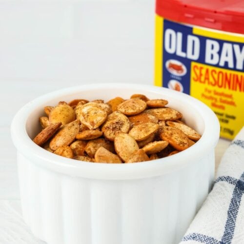 Old Bay seasoned pumpkin seeds in a white bowl in front of a container of Old Bay seasoning blend.