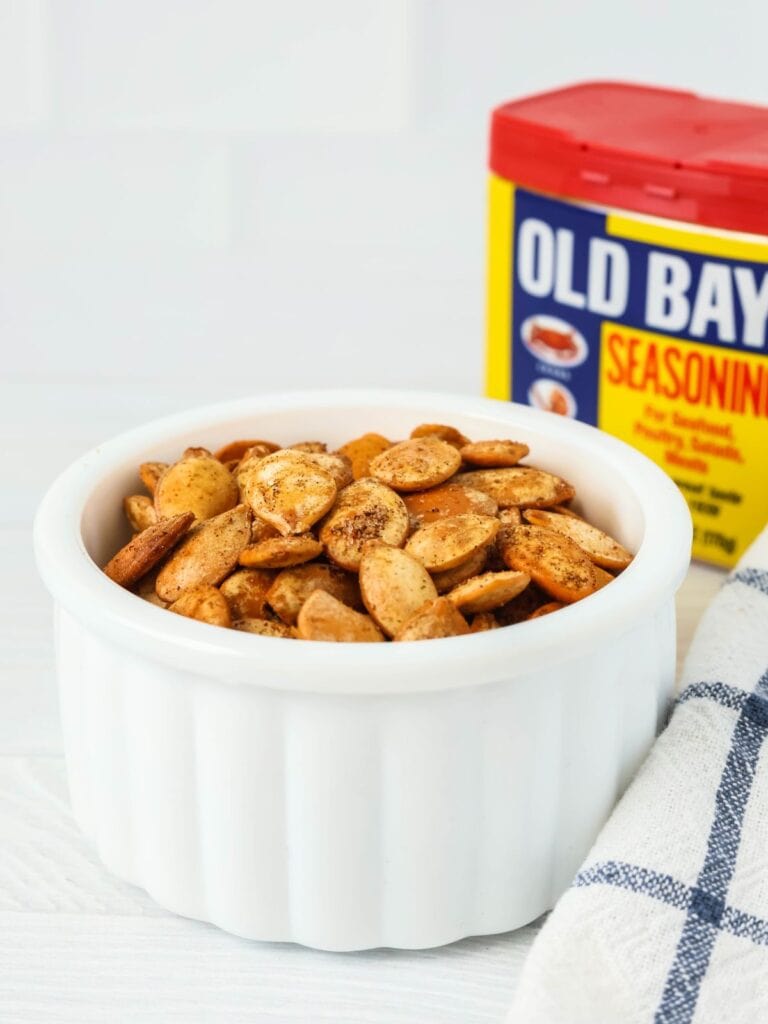 Old Bay seasoned pumpkin seeds in a white bowl in front of a container of Old Bay seasoning blend.