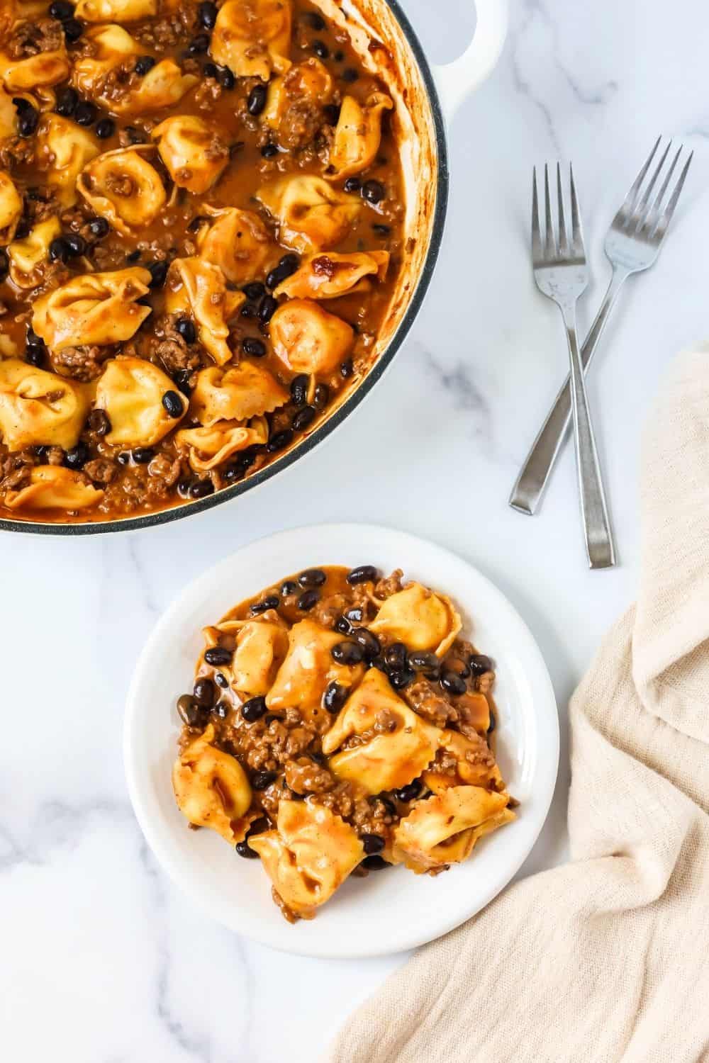overhead view of a plate of one pot enchilada tortellini, with the pan of remaining tortellini in the background. Two forks are next to the plate.