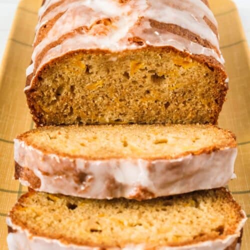 a loaf of homemade peach bread on a serving plate, with two slices cut from the loaf, showing the moist interior dotted with pieces of peaches.