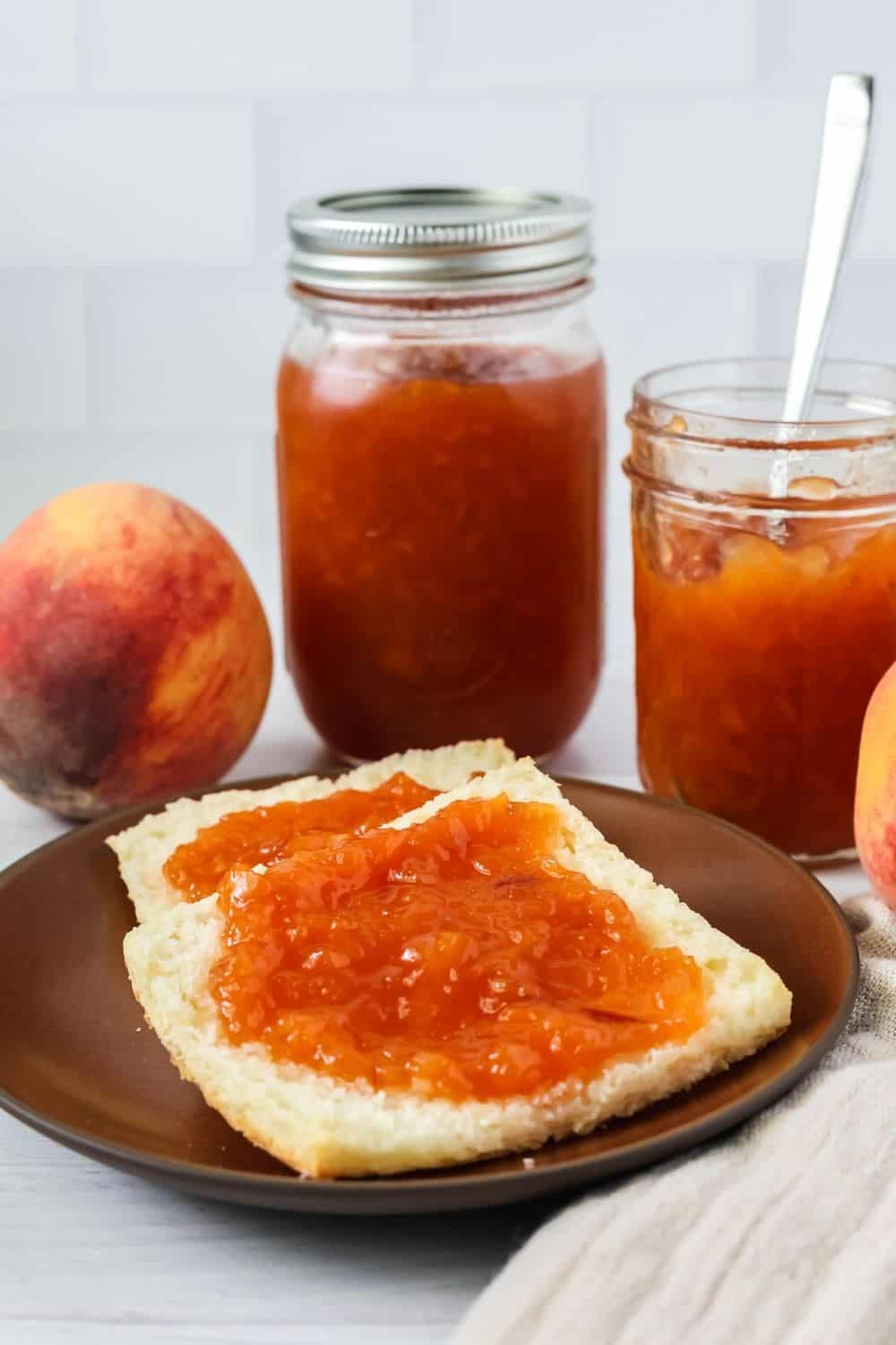 two halves of a biscuit are spread with peach jam made with the peels. A couple of jars of jam are in the background, along with a fresh whole peach.