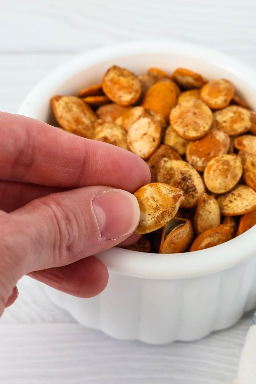 a woman's hand holds a pumpkin seed roasted with Old Bay seasoning in front of a bowl of seeds.
