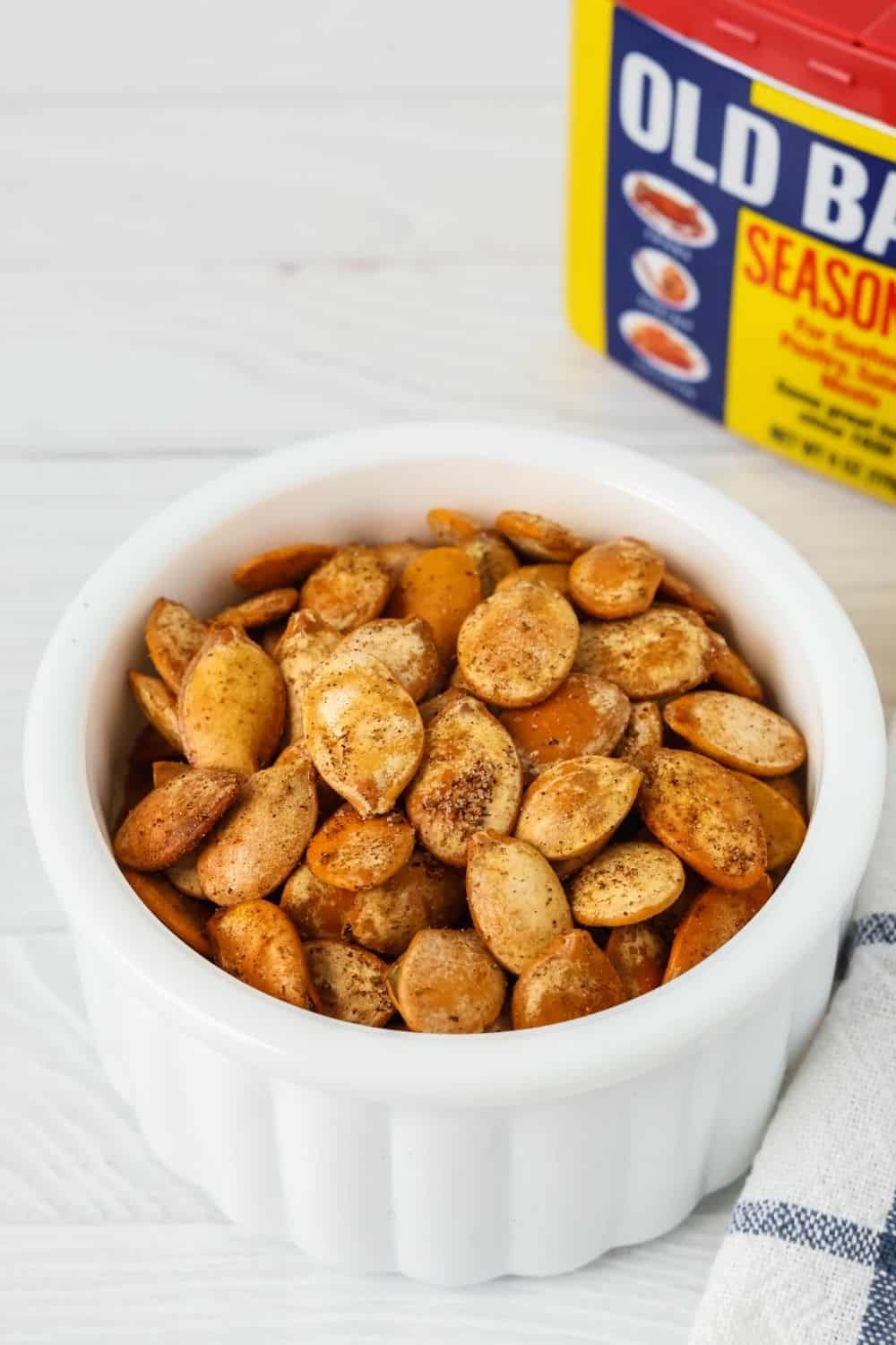 a white dish contains roasted pumpkin seeds seasoned with Old Bay seasoning. A container of Old Bay is in the background.