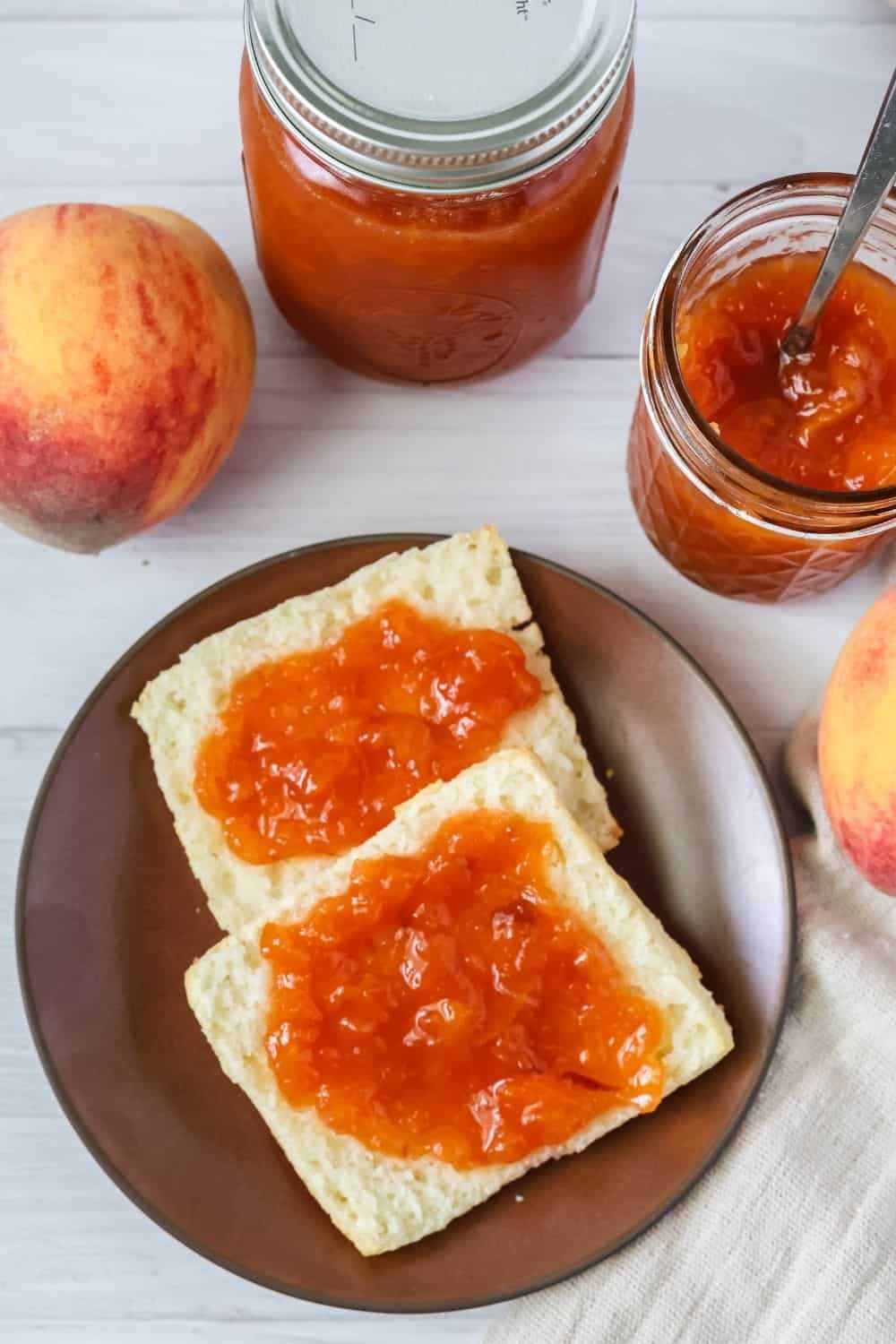 overhead view of biscuits topped with homemade southern peach jam, alongside fresh whole peaches and jars of jam.