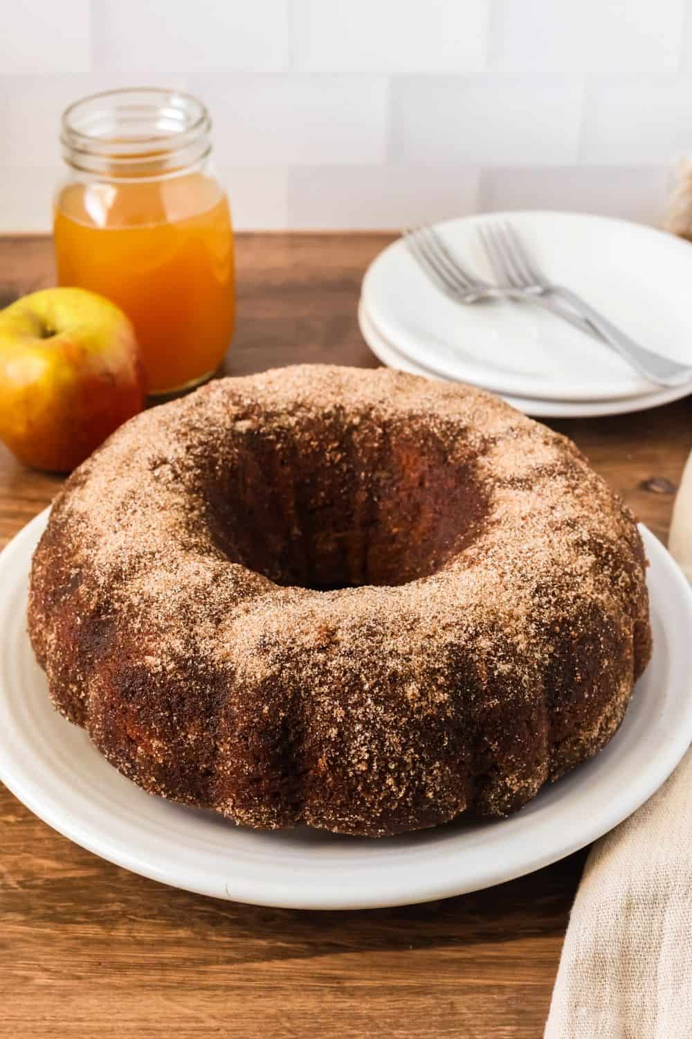 apple cider bundt cake made with spice cake mix is served on a white plate. Two small dessert plates and forks are in the background, along with a glass of apple cider and a fresh apple.