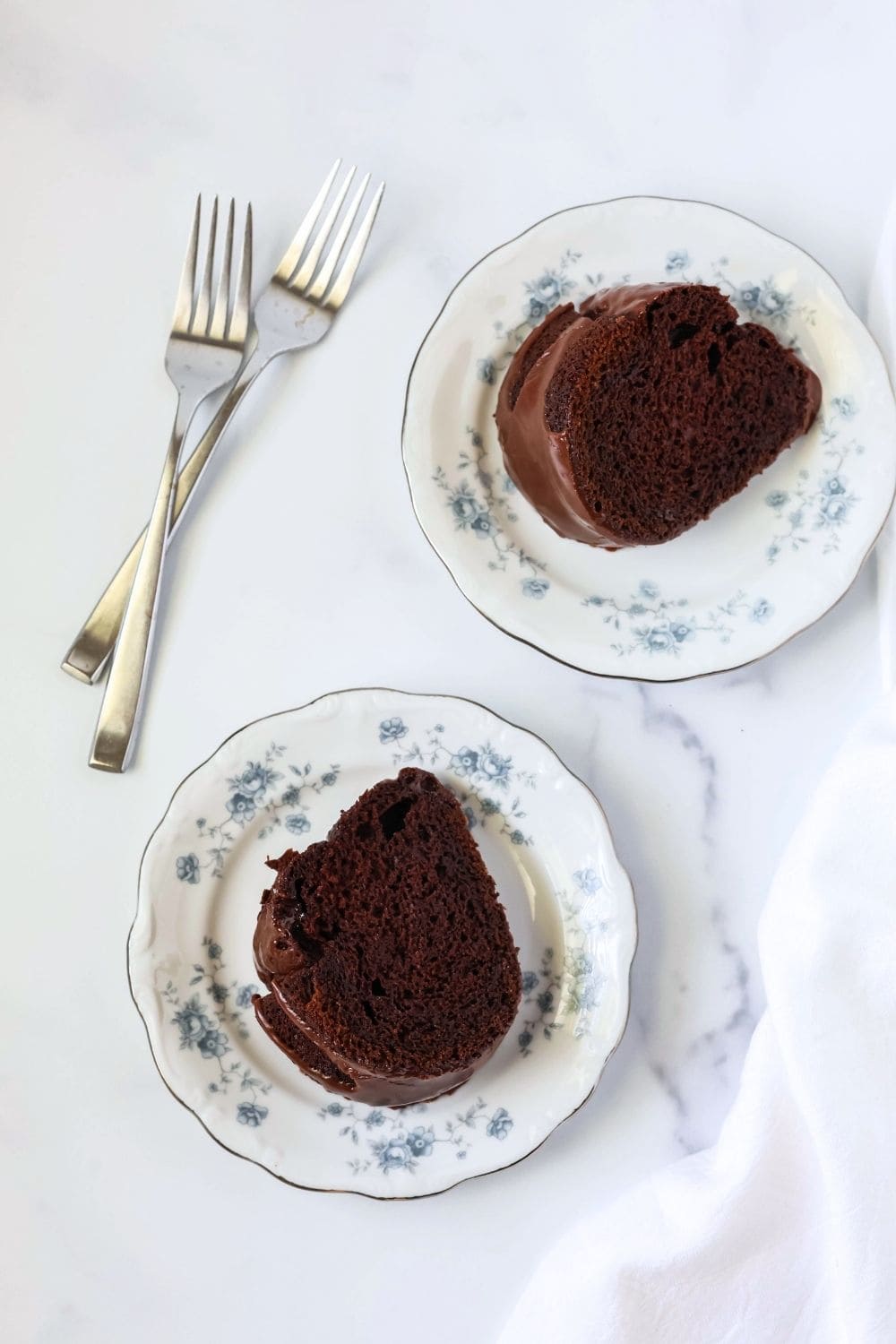 overhead view of two floral china plates serving slices of iced chocolate mayonnaise bundt cake, with two forks next to the plates.