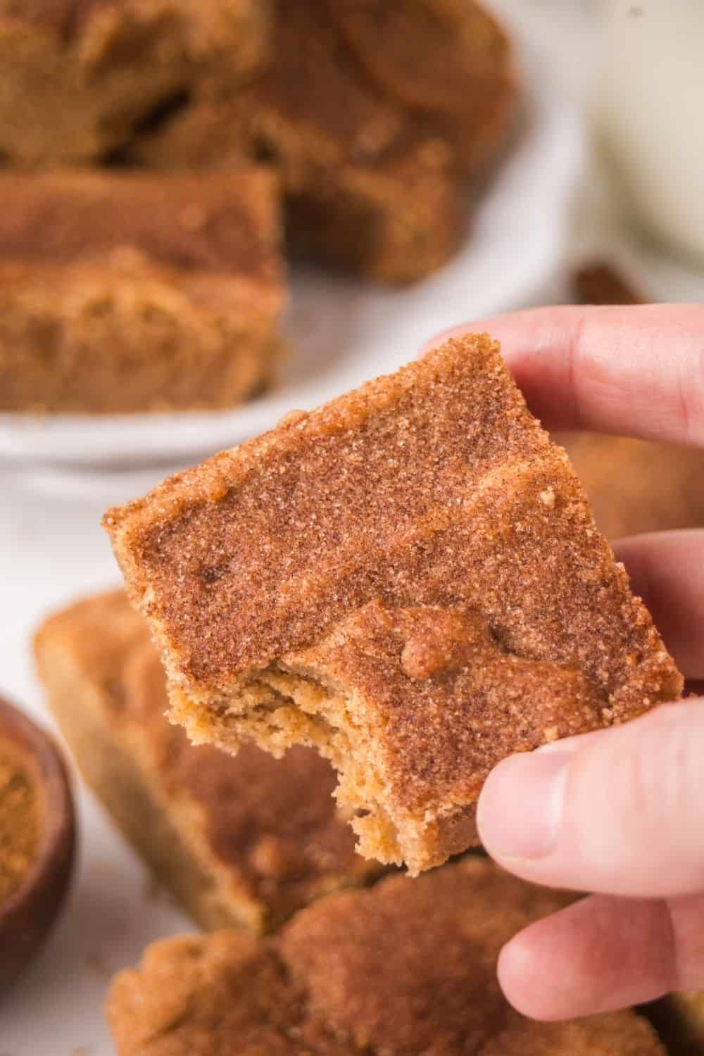 a hand holds a churro blondie bar square with a bite taken out of it.
