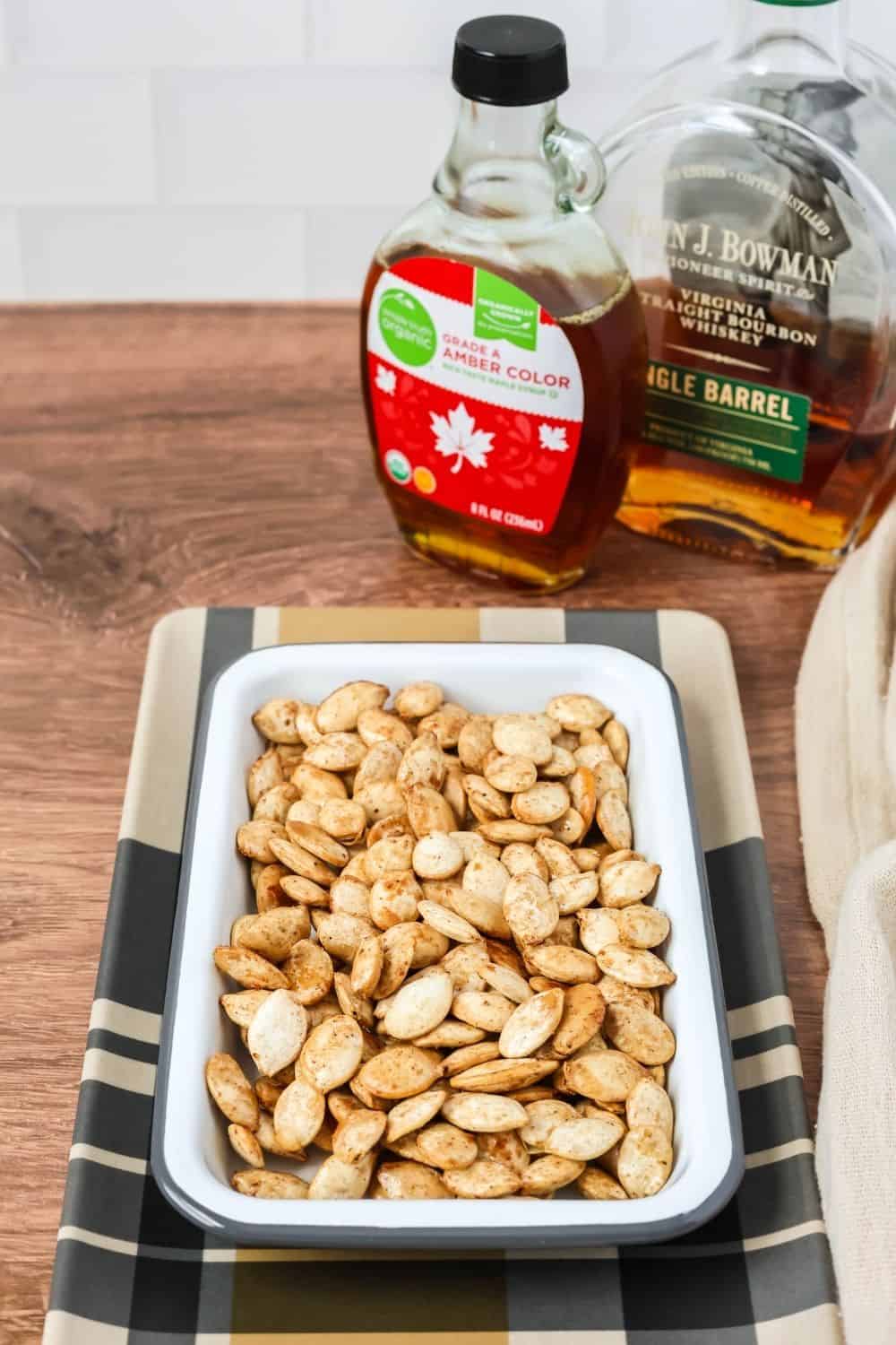 a white dish of maple bourbon roasted pumpkin seeds, with a bottle of maple syrup and a bottle of bourbon in the background.