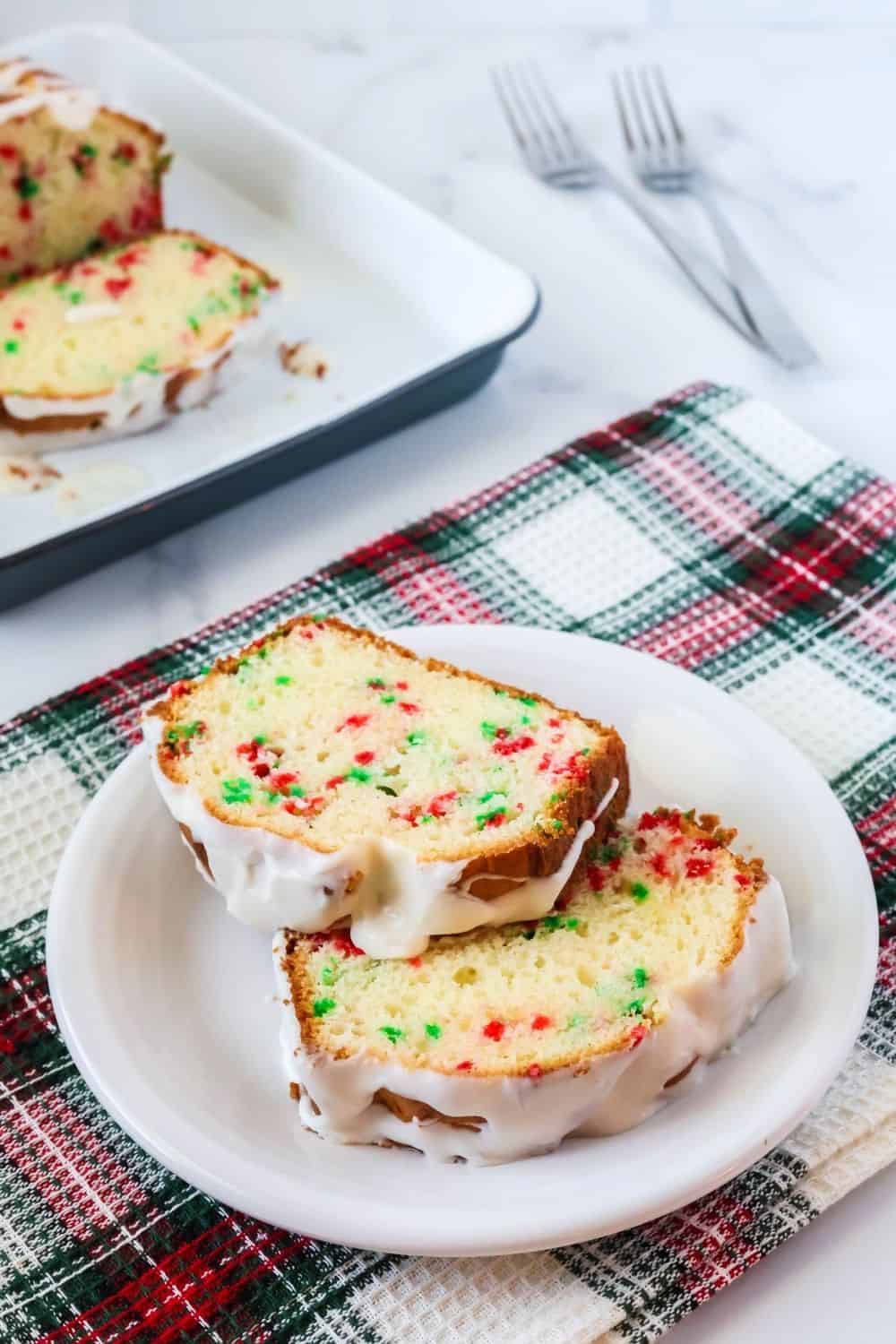 two slices of cake mix Christmas bread on a white plate, dotted with red and green sprinkles and topped with a drizzle of icing glaze. In the background is the remaining loaf of Christmas bread and two forks.