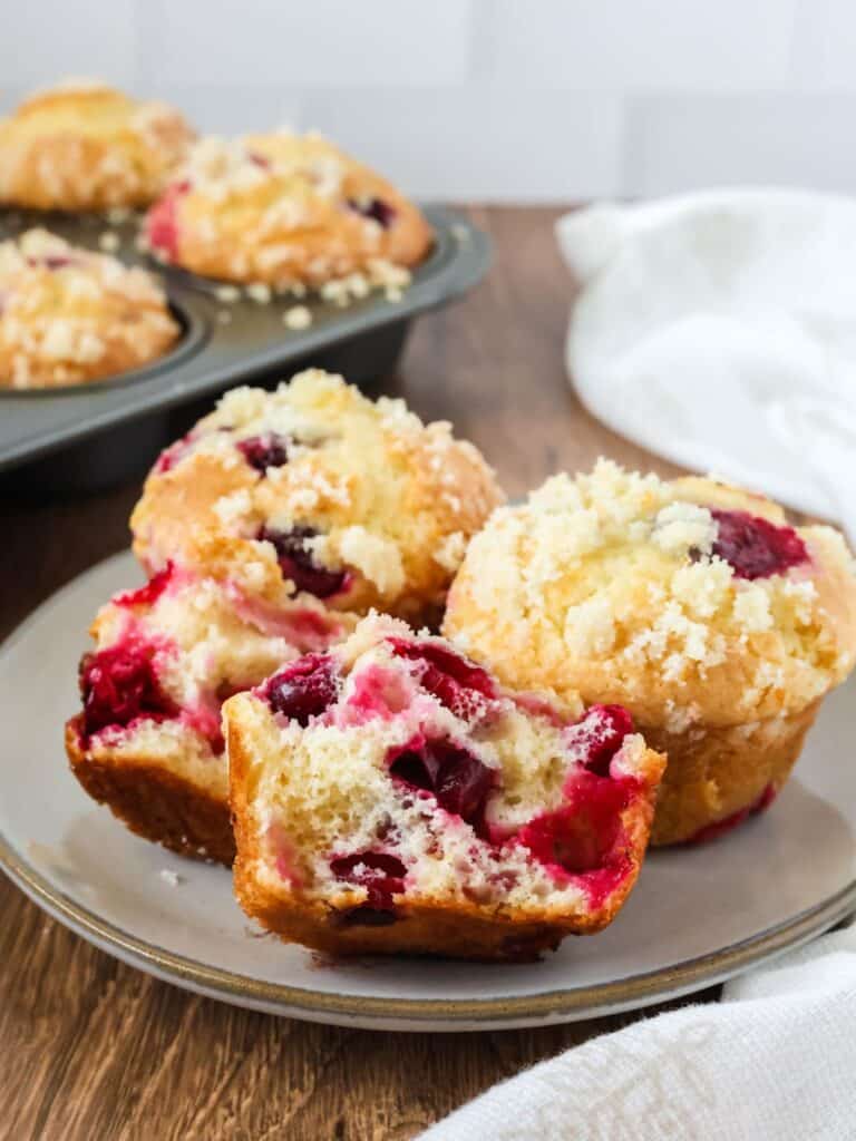 Cake mix cranberry orange muffins served on a plate, with more muffins in the pan in the background. One muffin is torn in half to show the juicy berries inside.