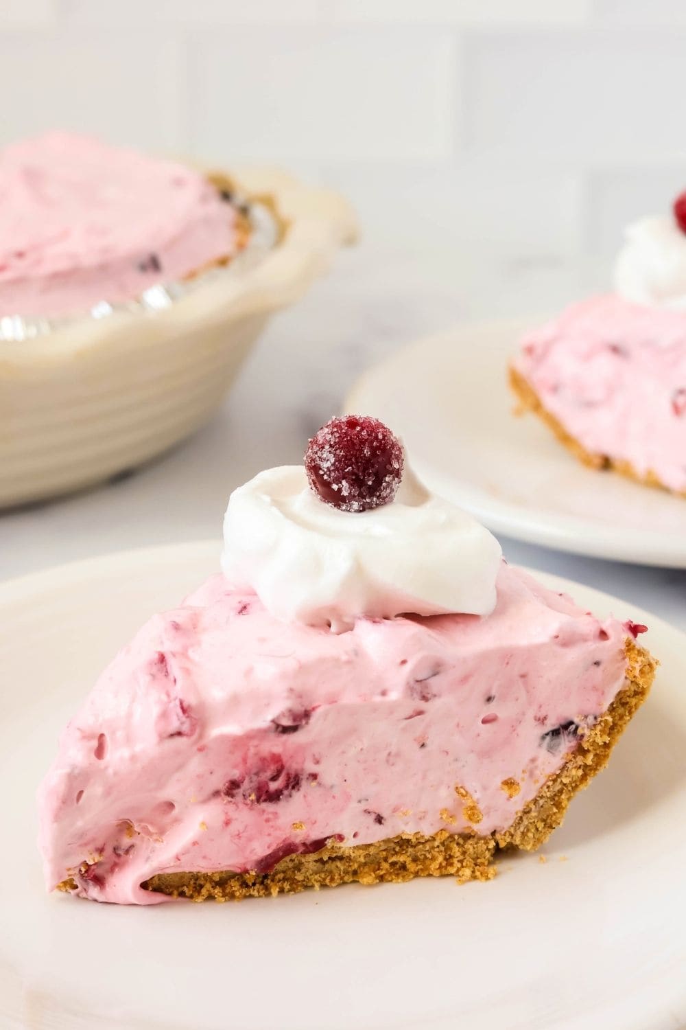 side view of a slice of no-bake cranberry Cool Whip pie on a white plate, garnished with whipped cream and a sugared cranberry. The remainder of the whole pie and another slice are in the background.
