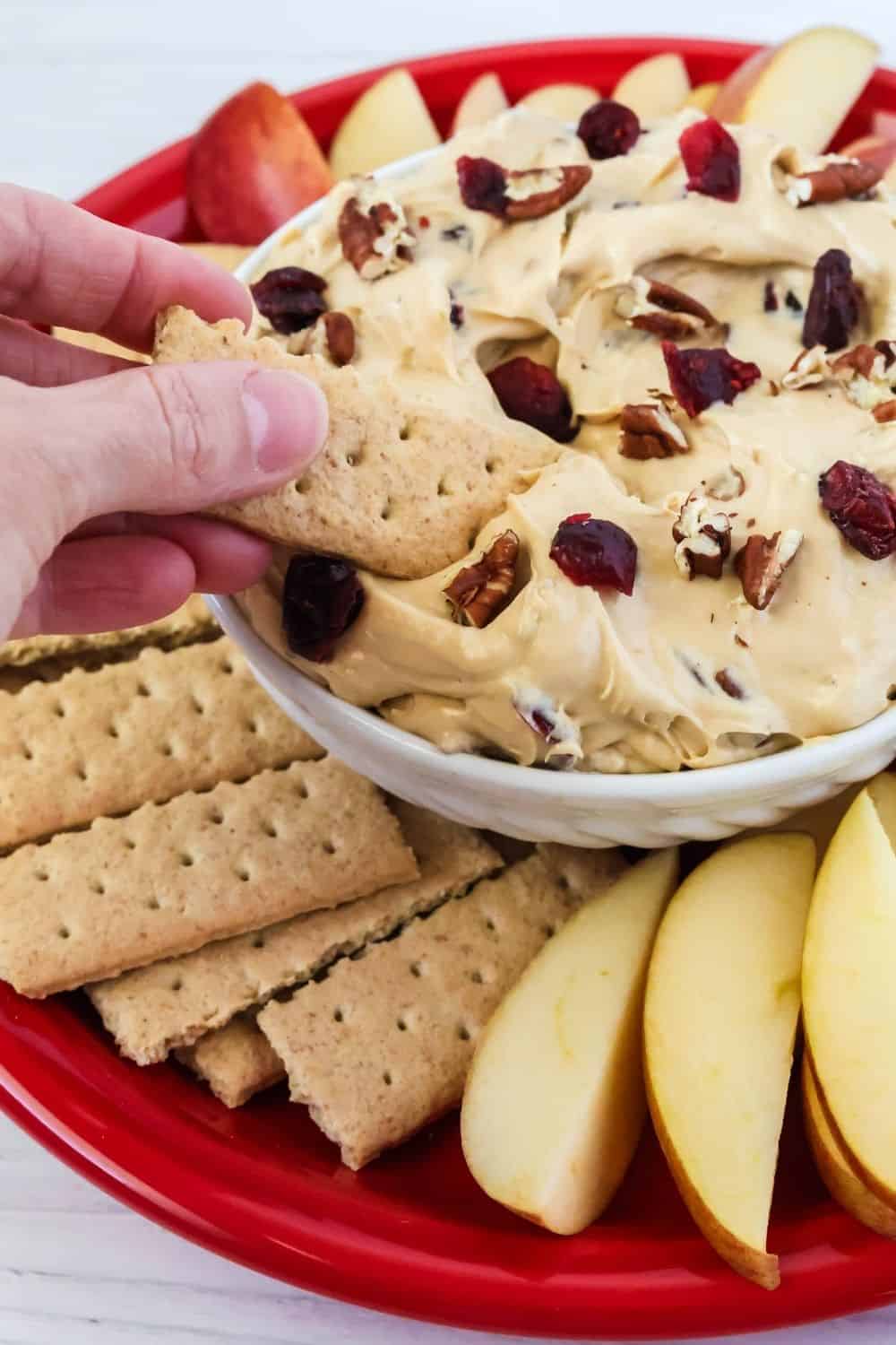 a woman's hand dips a graham cracker into a bowl of cranberry pecan fruit dip.