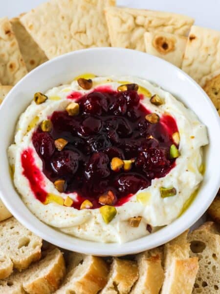 Holiday cranberry whipped ricotta dip topped with chopped pistachios, served in a white bowl along with bread dippers.