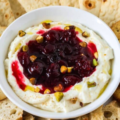 Holiday cranberry whipped ricotta dip topped with chopped pistachios, served in a white bowl along with bread dippers.
