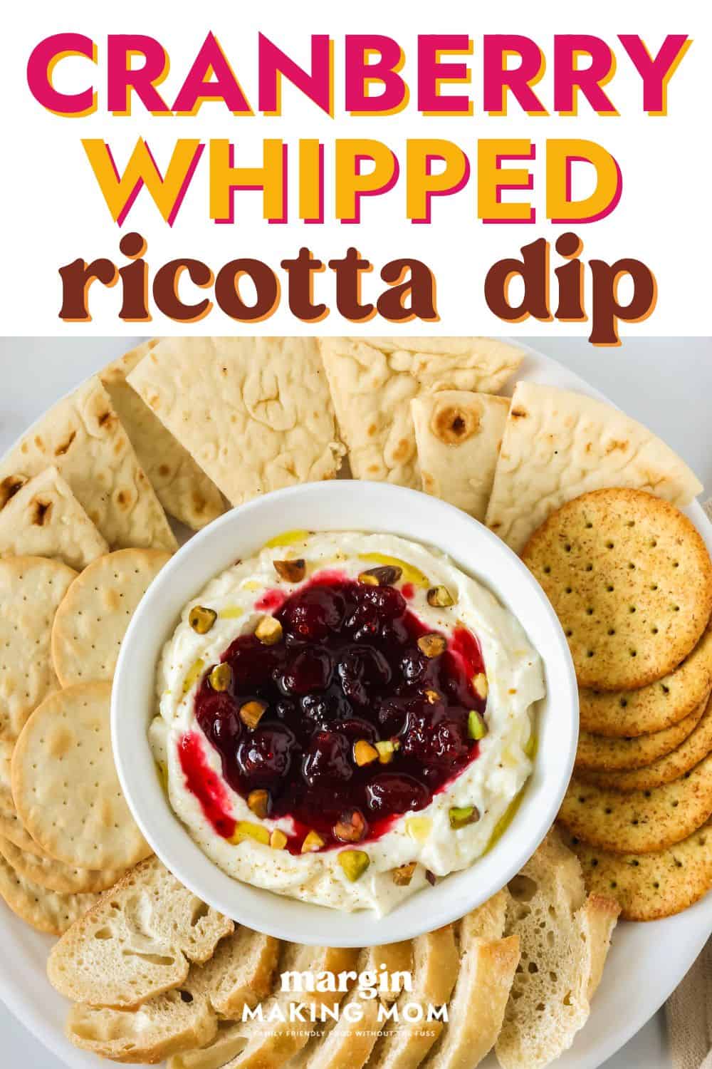 overhead view of a bowl of cranberry whipped ricotta dip on an appetizer platter, surrounded by crackers, bread slices, and pita triangles.