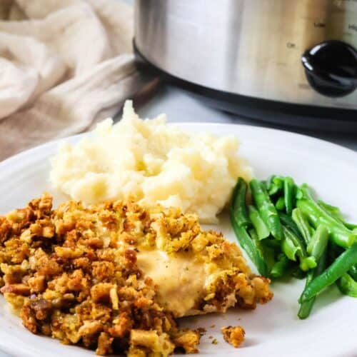 Crock Pot chicken and stuffing served on a white plate alongside mashed potatoes and green beans. A slow cooker is in the background.