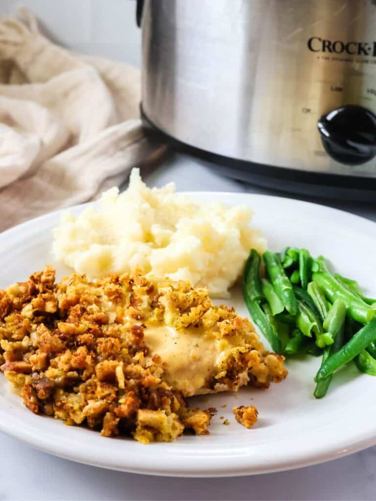 Crock Pot chicken and stuffing served on a white plate alongside mashed potatoes and green beans. A slow cooker is in the background.