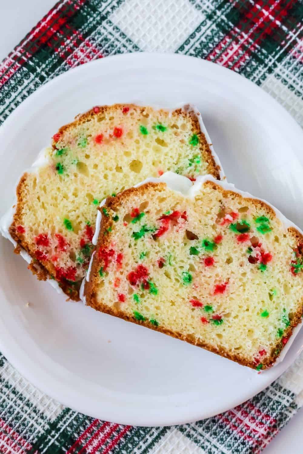 overhead view of two slices of Funfetti Christmas bread served on a white plate. The slices are dotted with red and green and topped with a white icing glaze.