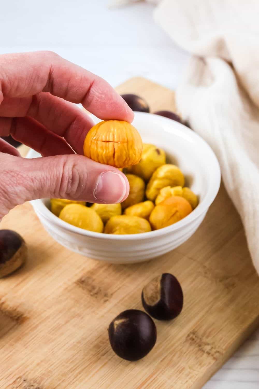 a woman's hand holds the meat of a roasted chestnut next to a bowl of more chestnuts.