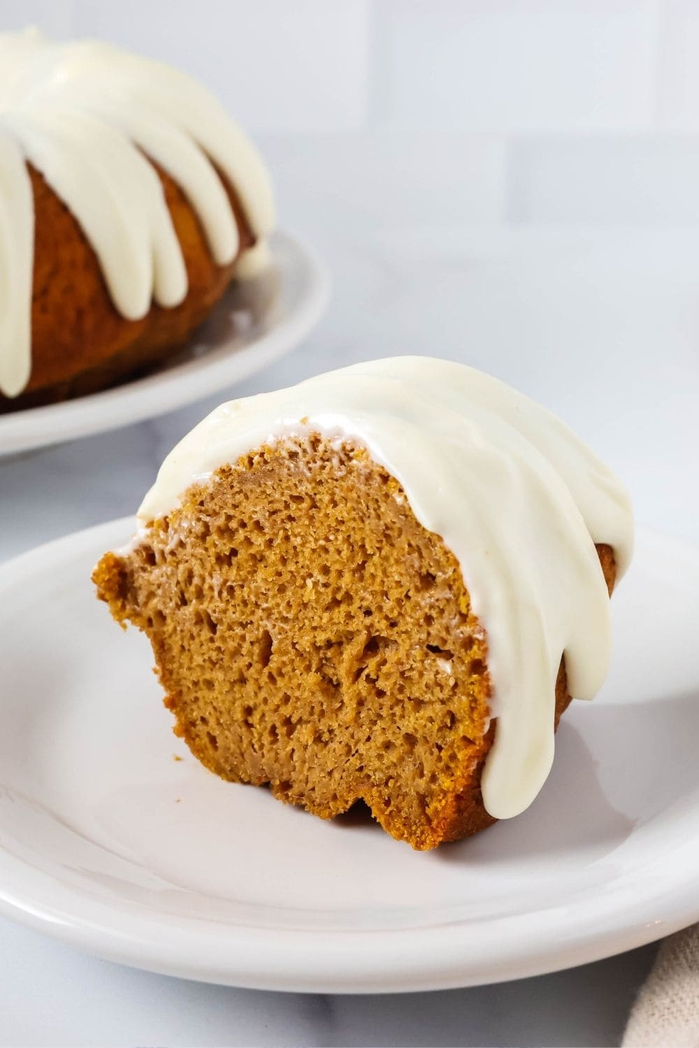 an upright slice of easy pumpkin bundt cake on a white plate, showing the moist interior and the drizzle of cream cheese glaze.