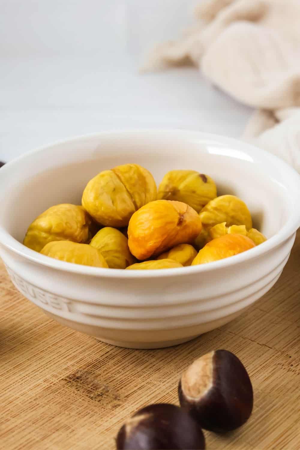 side view of holiday roasted chestnuts appetizer in a small white bowl. Two fresh chestnuts in the shell are in front of the bowl.