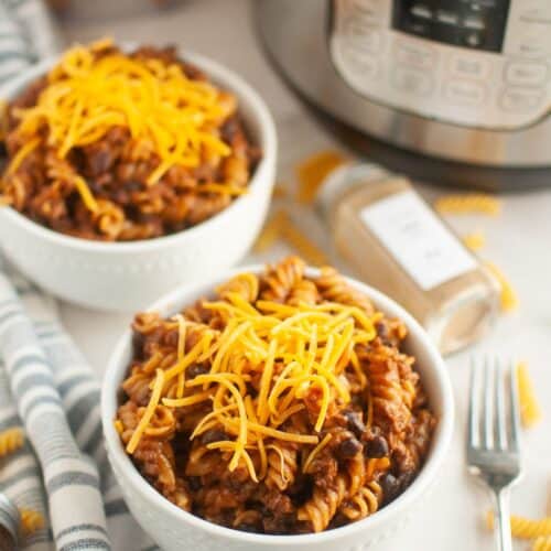 two white bowls of Instant Pot beef enchilada pasta garnished with shredded cheese. An Instant Pot pressure cooker is in the background.