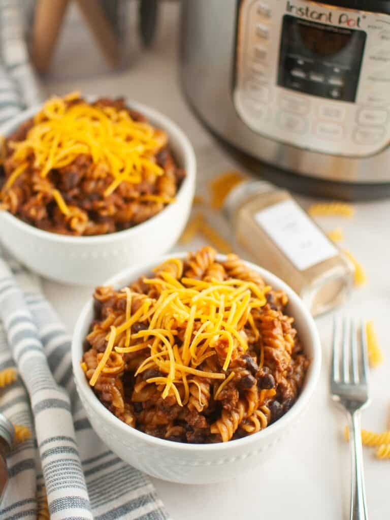 two white bowls of Instant Pot beef enchilada pasta garnished with shredded cheese. An Instant Pot pressure cooker is in the background.