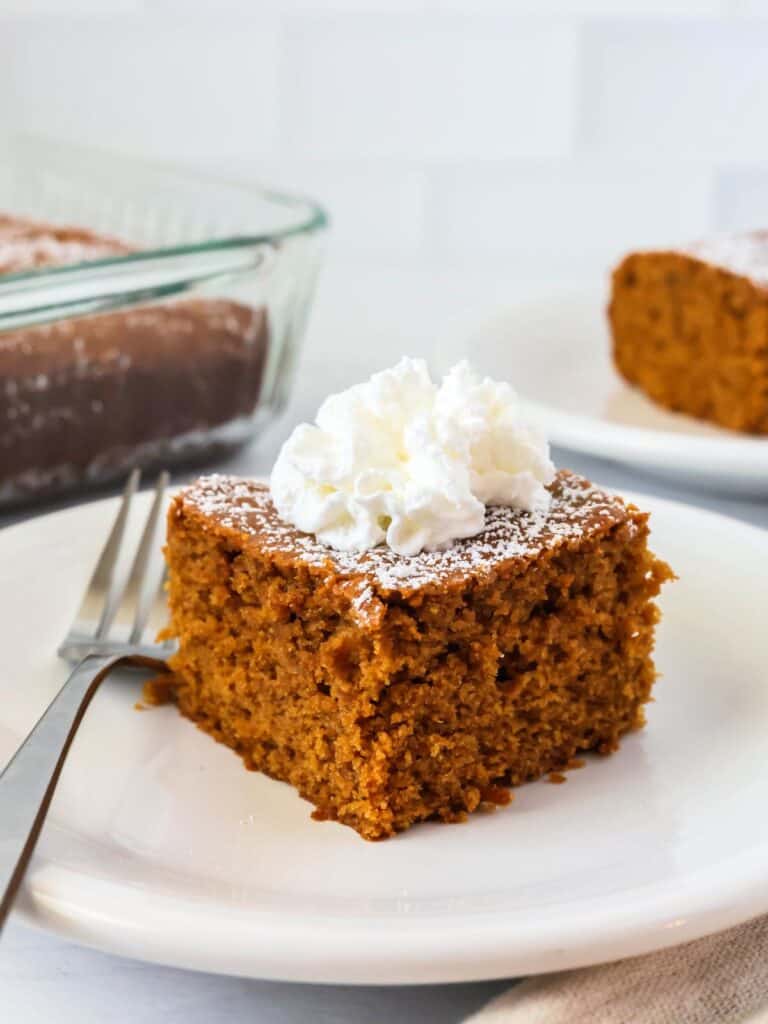 a slice of homemade molasses cake topped with whipped cream and served on a white plate, with a fork next to it.
