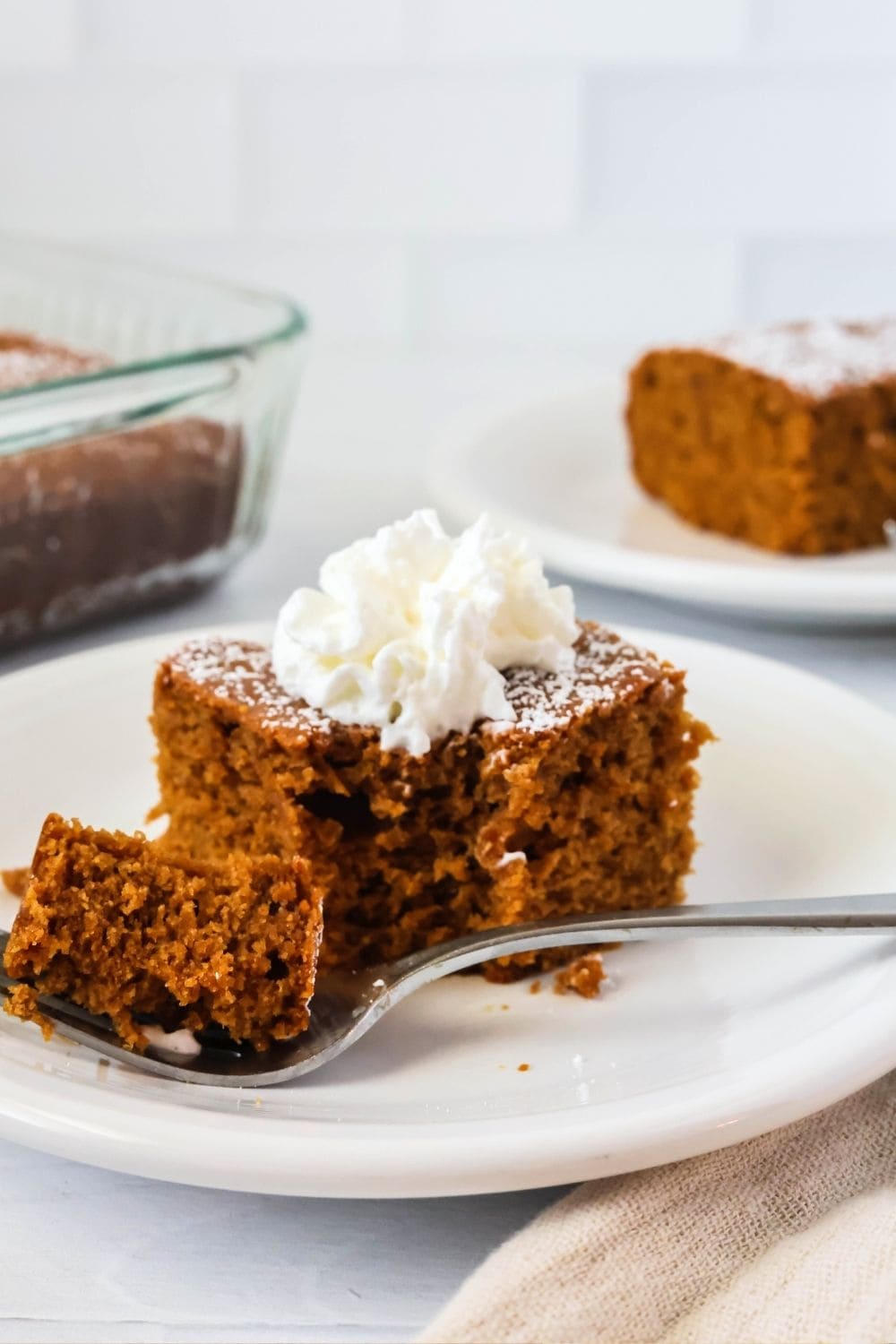 a slice of molasses cake topped with whipped cream on a white plate; a fork has cut a bite out of the cake and is resting on the plate alongside it.