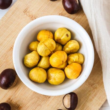 Roasted chestnuts served in a white bowl, their shells discarded so only the meat remains. Fresh chestnuts are surrounding the bowl.