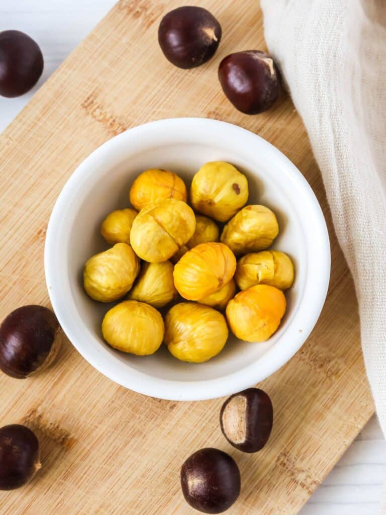 Roasted chestnuts served in a white bowl, their shells discarded so only the meat remains. Fresh chestnuts are surrounding the bowl.