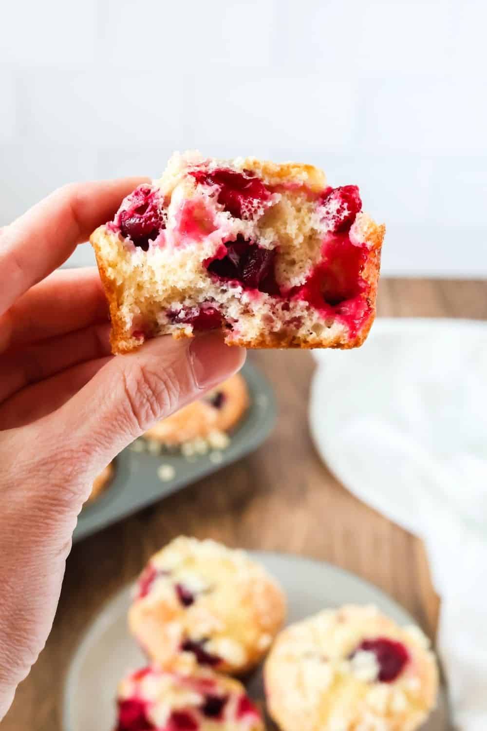 a hand holds half of a cranberry orange muffin made with cake mix. The remaining muffins from the batch are in the background.