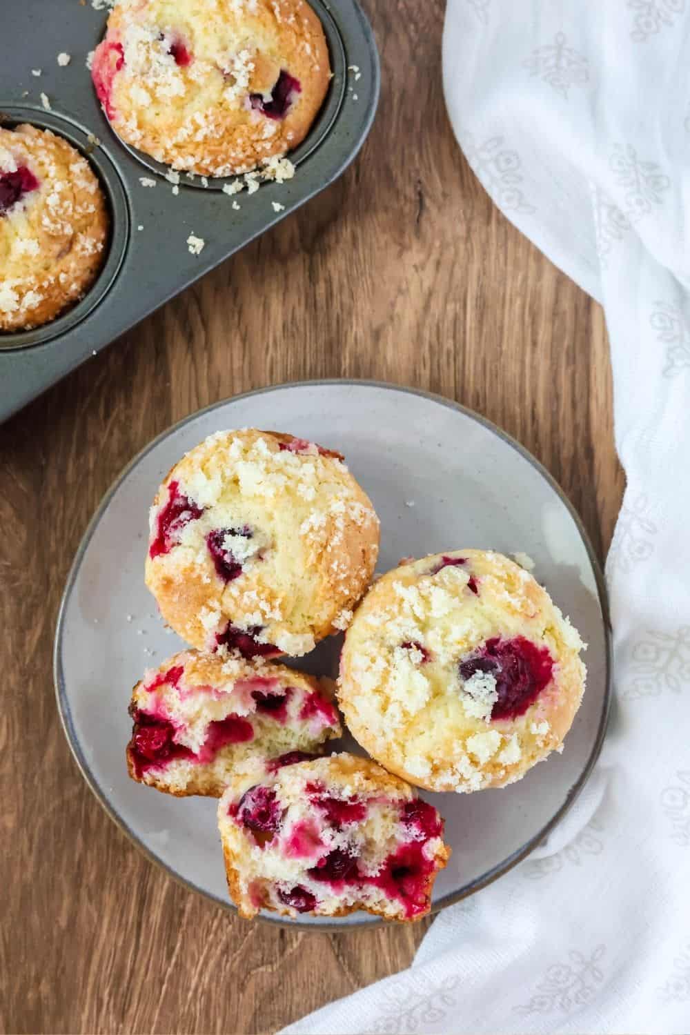 overhead view of easy cranberry orange muffins on an ivory-colored plate. One muffin has been torn in half to show all of the cranberries inside. A muffin pan is in the background.
