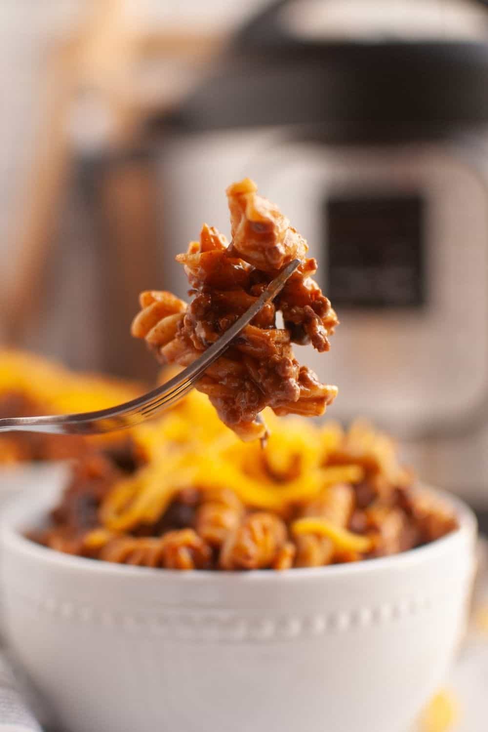 side view of a fork holding a bite of beef enchilada pasta. A pasta bowl and Instant Pot are in the background.