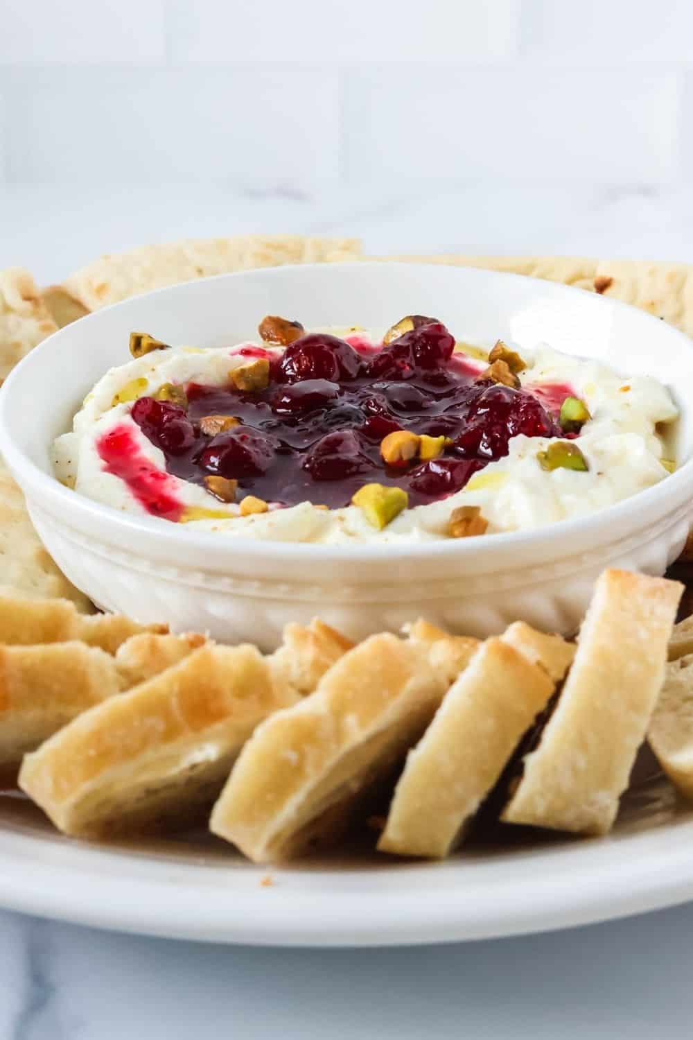 close-up view of cranberry whipped ricotta topped with pistachio nuts. Sliced baguette is next to the dip bowl.