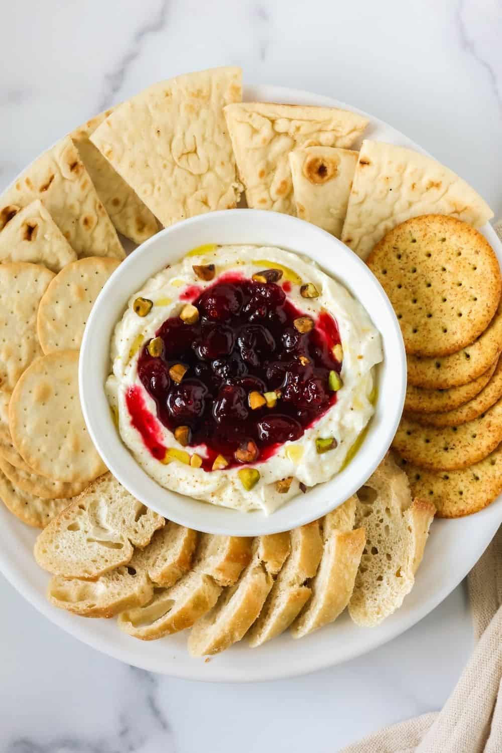 overhead view of a white bowl of cranberry whipped ricotta for the holidays, surrounded by crackers, pita wedges, and baguette slices.